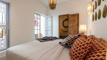 Une chambre avec un grand lit, une fenêtre ornée de barreaux en fer forgé, une armoire en bois et un luminaire fixé au mur.