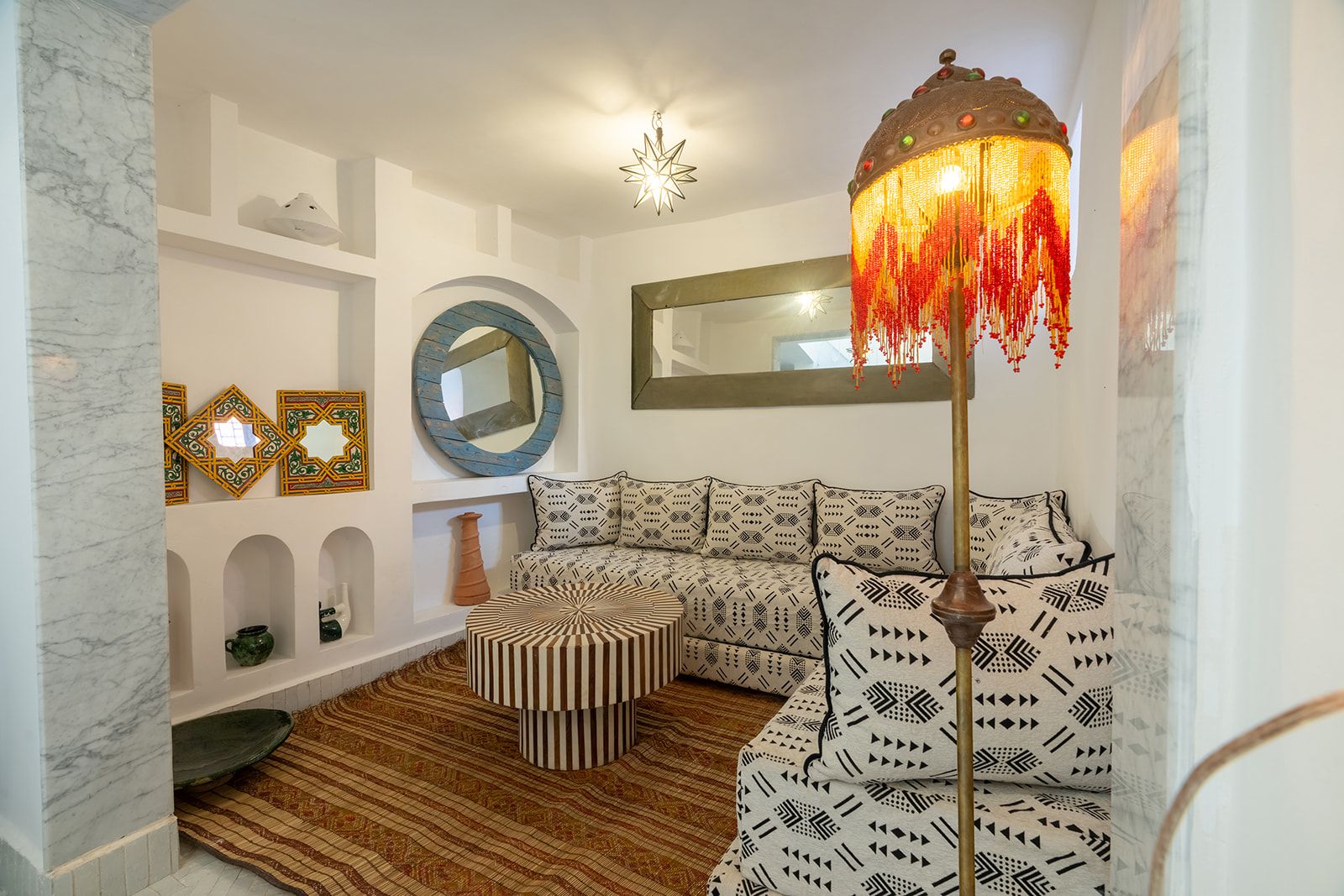 Un salon moderne avec un canapé d’angle, un pouf à motifs, une lampe de sol décorative et un grand miroir.