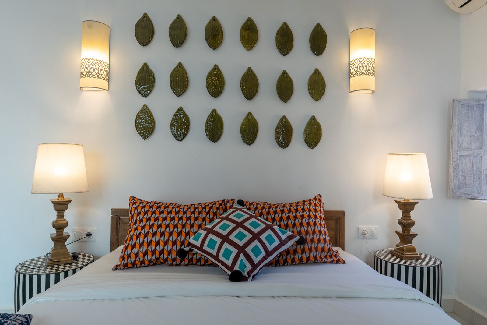 Een slaapkamer met een bed, decoratieve kussens met een patroon, twee bedlampen en decoratieve objecten aan de muur.