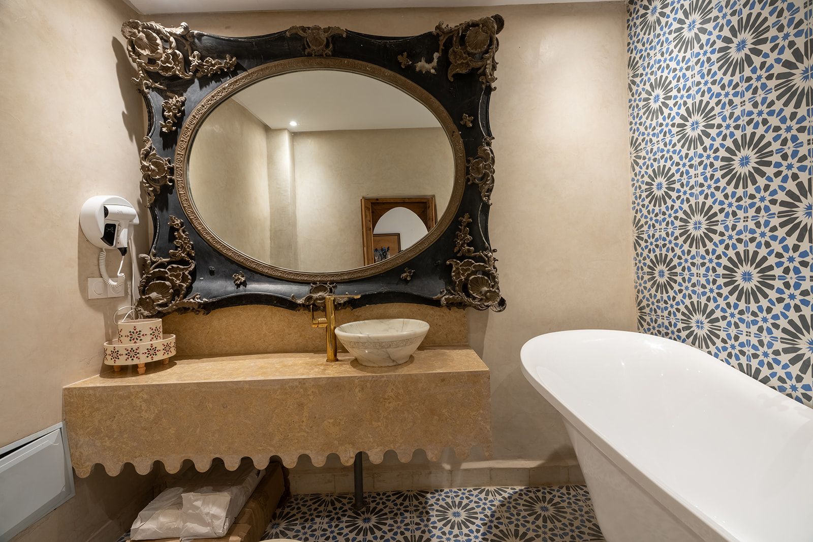 Une salle de bain dotée d’un grand miroir décoratif, d’un lavabo blanc avec un robinet doré et d’une baignoire blanche.