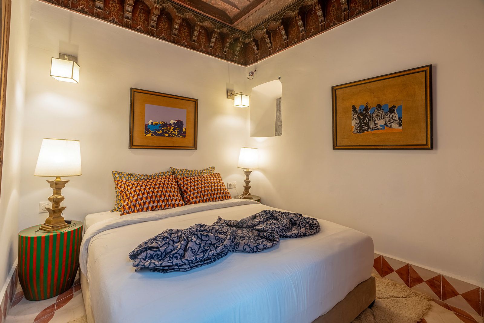 Une chambre d’hôtel avec un lit, deux lampes de chevet, des coussins à motifs, un tableau encadré au-dessus du lit et un tapis à motifs sur le sol.