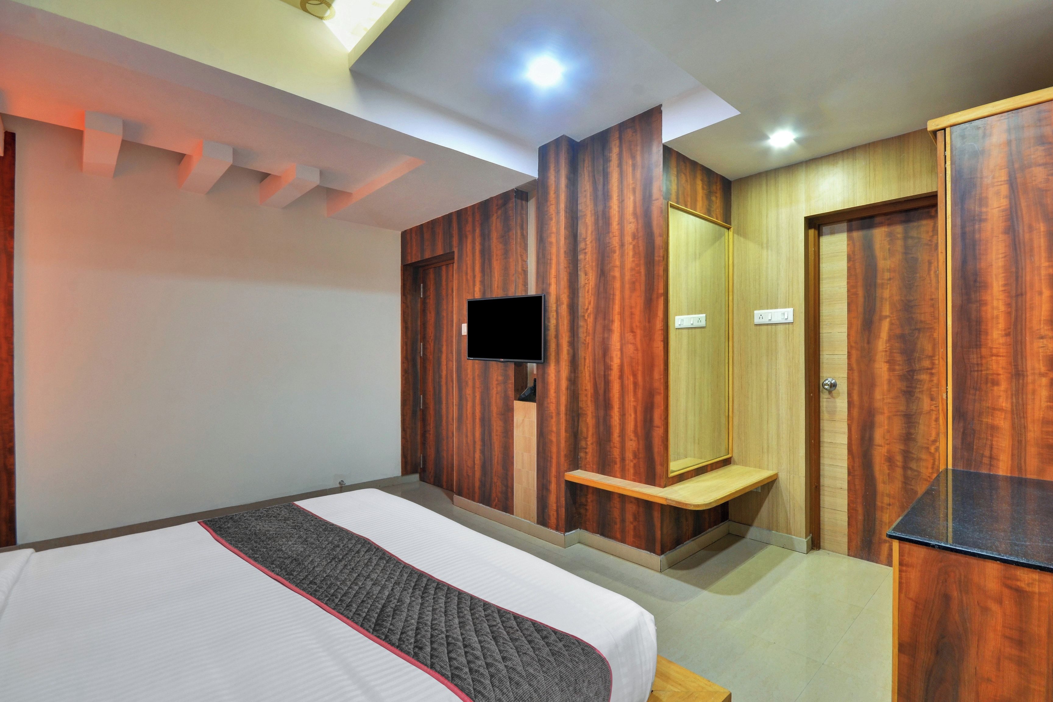 A hotel room with a bed, a flat-screen TV mounted on the wall, a wooden wardrobe, and a small wooden shelf.