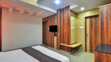 A hotel room with a bed, a flat-screen TV mounted on the wall, a wooden wardrobe, and a small wooden shelf.