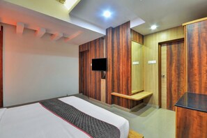 A hotel room with a bed, a flat-screen TV mounted on the wall, a wooden wardrobe, and a small wooden shelf.