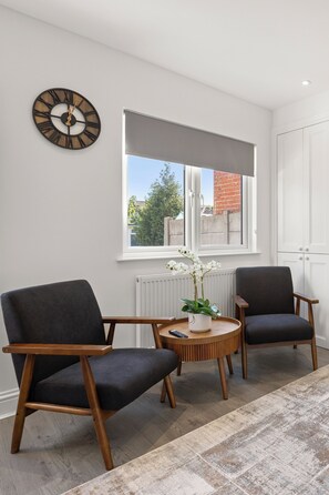 A room with two armchairs, a round table, a wall clock, a window with a view, and a radiator. - Modern London Home | 4BR | 2 bath & Parking (Ilford)