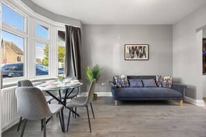 A modern living room with a dining area, grey walls, a blue sofa, and a large window with curtains. - Modern London Home | 4BR | 2 bath & Parking (Ilford)