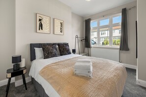 A neatly made bed with a white and beige blanket, a black headboard, and two framed artworks on the wall. - Modern London Home | 4BR | 2 bath & Parking (Ilford)