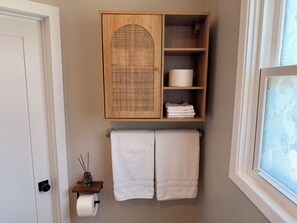 A bathroom with a wooden cabinet, two towels, a toilet paper holder, and a window with a patterned curtain. - South Park’s Better Half (San Diego)