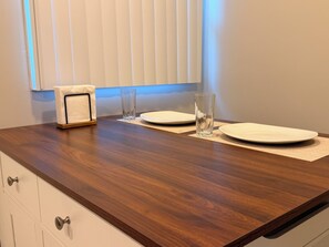 A wooden dining table with two plates and two glasses, a napkin holder, and a window with blinds. - South Park’s Better Half (San Diego)