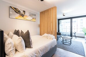 A modern hotel room with a large bed, a painting on the wall, a wooden wardrobe, a balcony with a view of buildings, and a small round table. - Newlands Peak Studio with AC and gym (Cape Town)