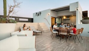 A modern outdoor dining area with a stone floor, surrounded by white furniture and a cactus plant.