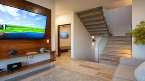 A modern living room with a flat-screen TV, wooden entertainment unit, and a staircase with glass railings.