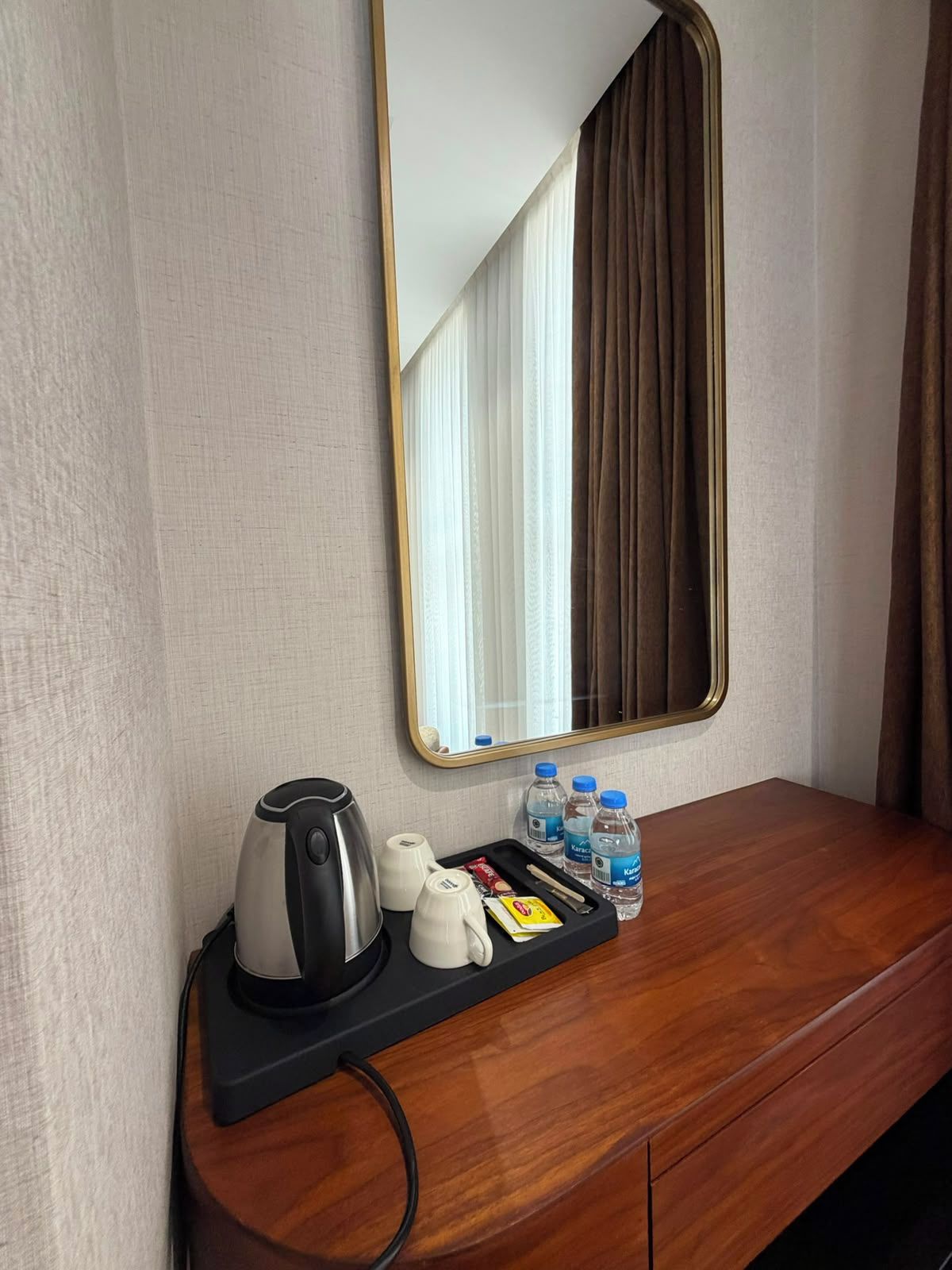 Habitación de hotel con una mesita de noche de madera, una bandeja negra con tetera, tazas y agua embotellada.