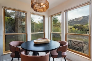 Un comedor con una mesa redonda y cuatro sillas, ventanas amplias con vistas a árboles y una montaña, y una lámpara de techo de fibra natural.