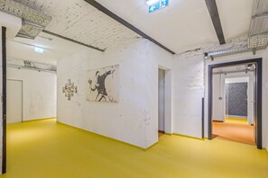 Ein Flur mit gelbem Boden, weißen Wänden und einem großen schwarz-weißen Wandgemälde. Rechts befinden sich Türen und die Decke zeigt freiliegende Balken und Leuchten.