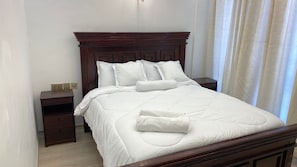 A neatly made bed with white linens and pillows, a wooden headboard, and a bedside table with a lamp. - COZY MODERN 1-BEDROOM IN WESTLANDS NEAR GTC AND MALLS (Nairobi)
