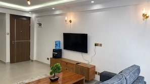 A modern living room with a flat-screen TV mounted on the wall, a wooden coffee table with a small plant, a grey sofa, and a water dispenser on a cabinet. - COZY MODERN 1-BEDROOM IN WESTLANDS NEAR GTC AND MALLS (Nairobi)