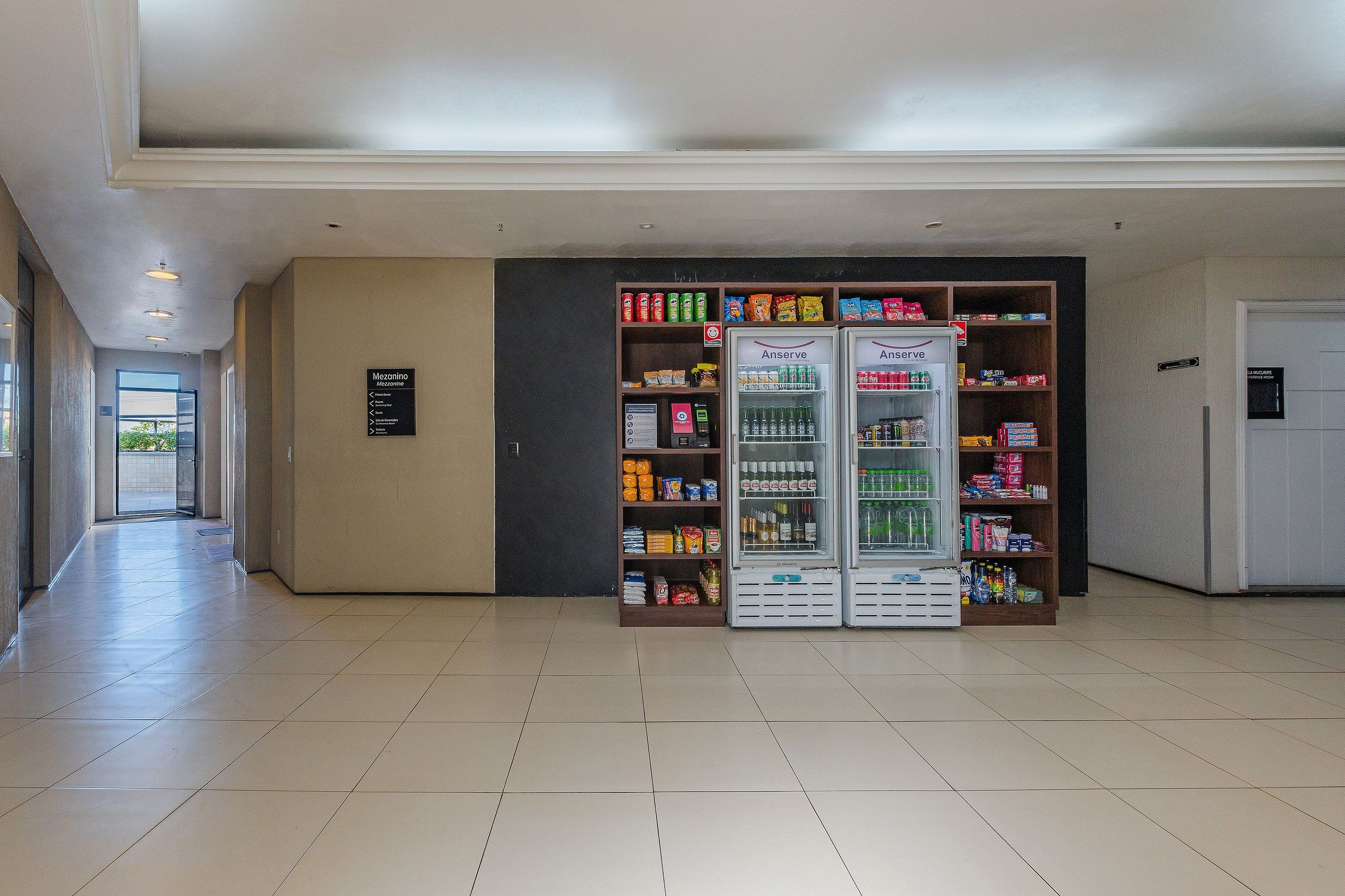 Corredor com máquinas de venda automática repletas de lanches e bebidas, uma placa de sinalização e uma porta para um quarto.