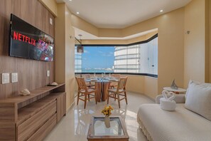 A modern living room with a flat-screen TV, wooden furniture, a dining area, and a view of the city through large windows. - Golden Flat - Seaside View Flat by Carpediem (Fortaleza)