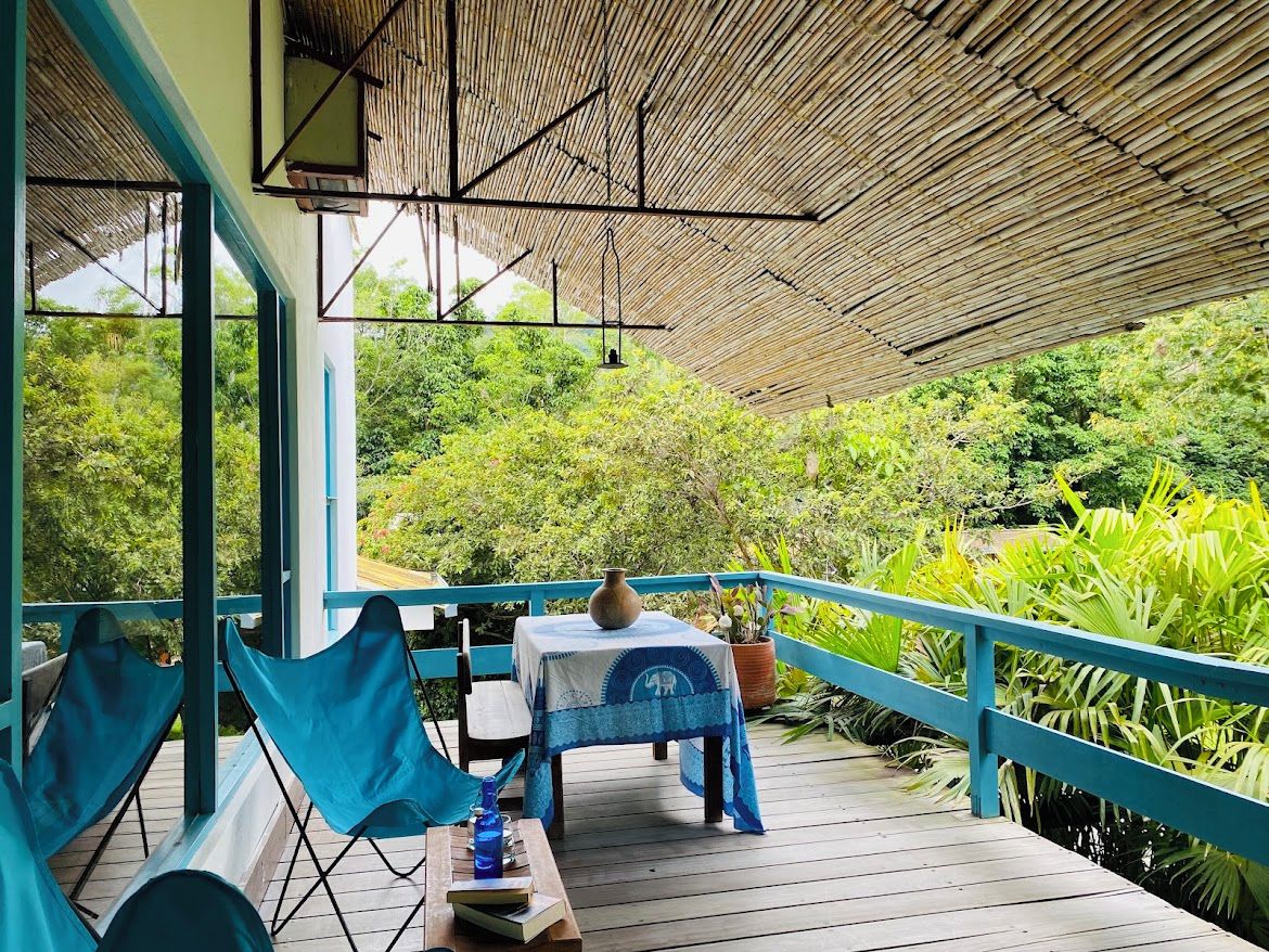 A covered outdoor area with a table and chairs, surrounded by lush greenery.