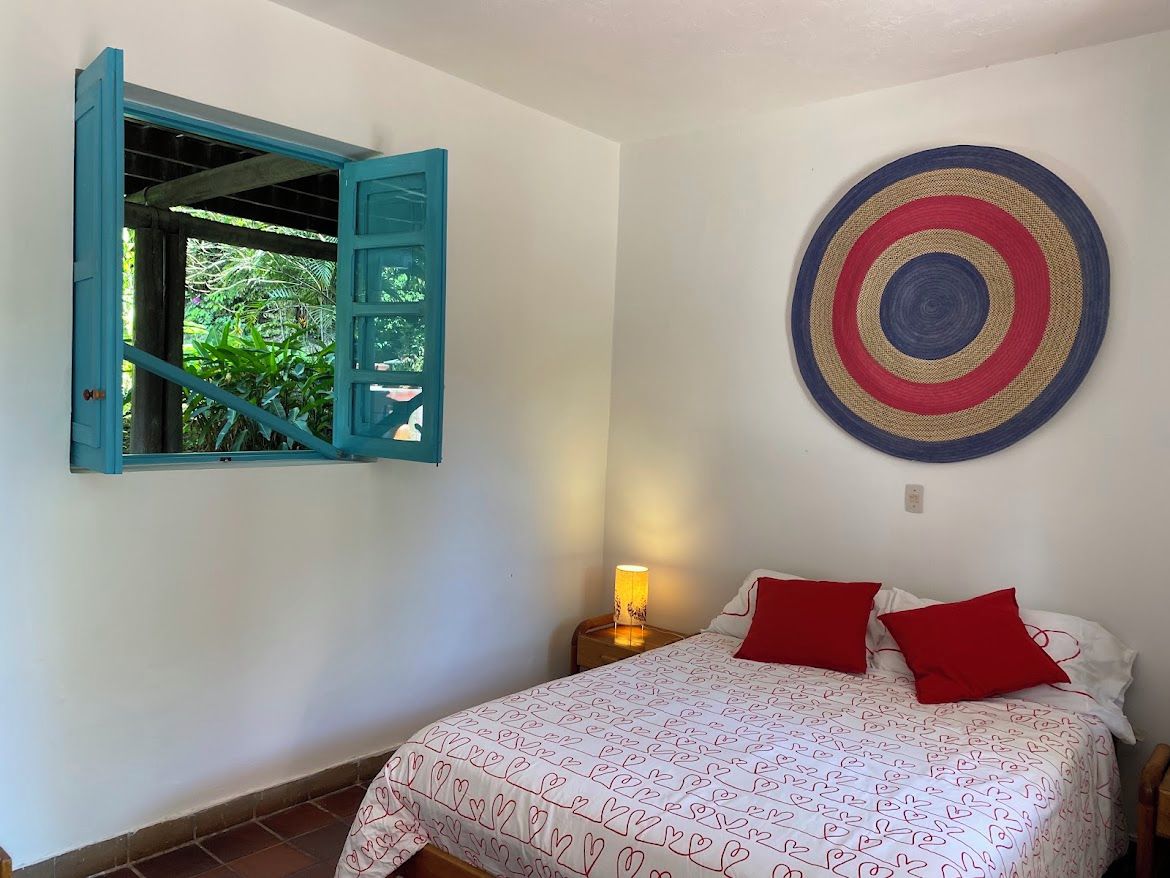 A bedroom with a bed, a wall-mounted circular artwork, a window with blue shutters, and a nightstand with a lamp.
