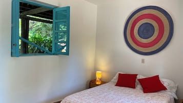 A bedroom with a bed, a wall-mounted circular artwork, a window with blue shutters, and a nightstand with a lamp.