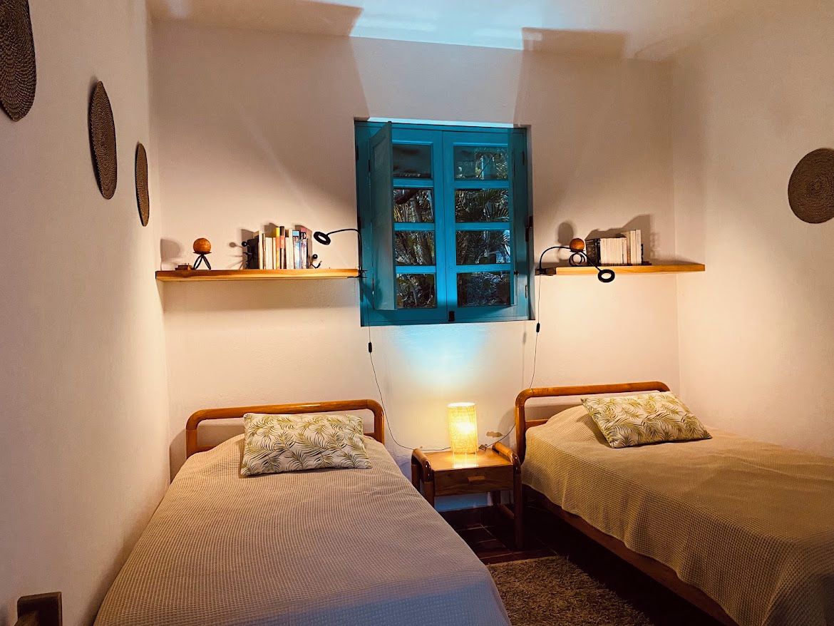 Une petite chambre à coucher accueillante, avec deux lits simples, une table de chevet avec une lampe et une fenêtre dotée d’un volet bleu.