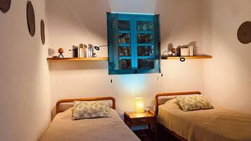 Une petite chambre à coucher accueillante, avec deux lits simples, une table de chevet avec une lampe et une fenêtre dotée d’un volet bleu.