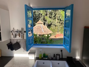A kitchen with a window view of a thatched-roof structure and trees.