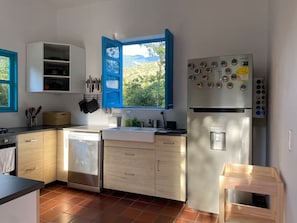 A modern kitchen with a dishwasher, a refrigerator with a water dispenser, and a window with blue shutters.