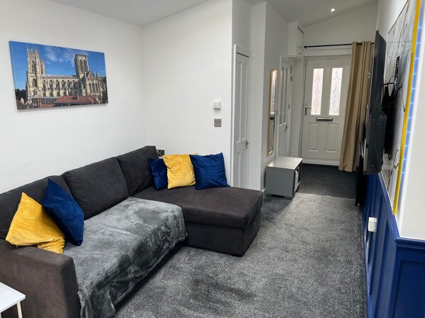 A modern living room with a sectional sofa, a flat-screen TV, and a painting of a cathedral. - House Sleeps 4 - Private Garden - Bbq (York)