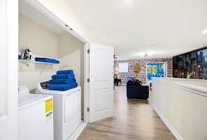 Zona de bugaderia amb rentadora i assecadora, espai per plegar roba amb tovalloles blaves, i habitació amb paret de pedra i quadre d'una escena urbana.