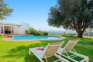 A swimming pool with a view of the countryside, two lounge chairs, and a large tree. - Raithbury Hill House (Stellenbosch)