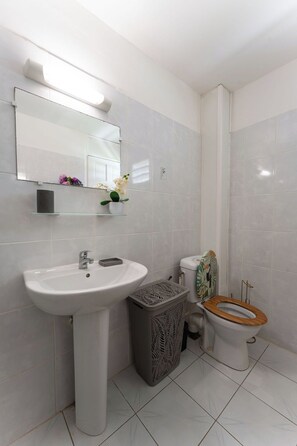 A bathroom with a pedestal sink, a toilet with a wooden seat, and a trash bin. - Air-conditioned studio with a terrace and pool, 5 minutes from the beaches of Sainte-Anne (Sainte-Anne)