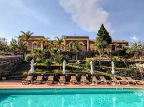 A swimming pool with lounge chairs and umbrellas, a stone staircase, and a building with arched windows and a tiled roof.
