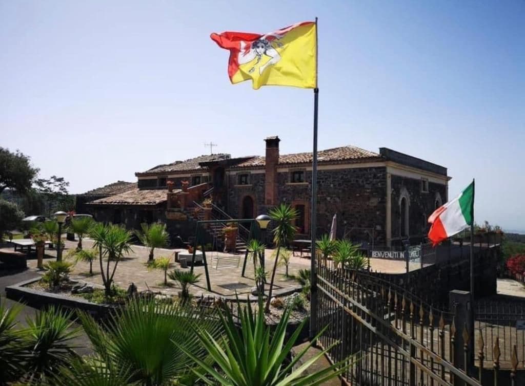 Un bâtiment à la façade en pierre, un mât portant un drapeau et une cour entourée d’une clôture, agrémentée de palmiers.