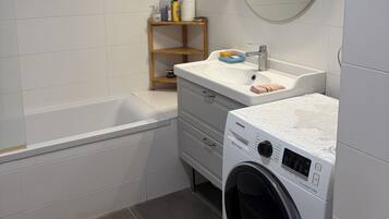 Een badkamer met een bad, een wasmachine, een wastafel met spiegel en een plank met toiletartikelen.