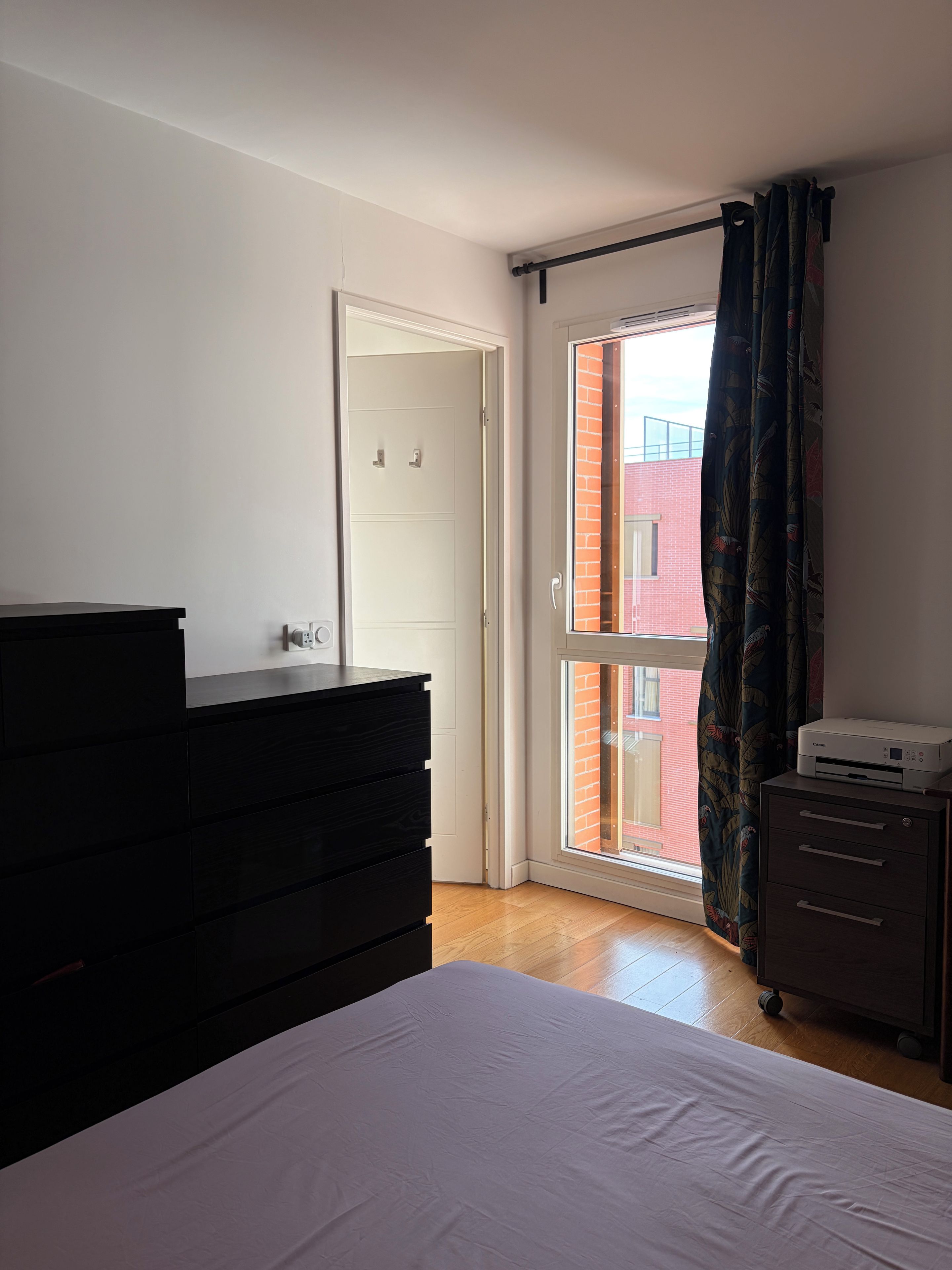 Een slaapkamer met een bed, een dressoir, een printer en een schuifpui naar een balkon.