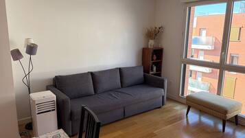 A modern living room with a grey sofa, a small table, a chair, a bookshelf, and a large window with a view of buildings.