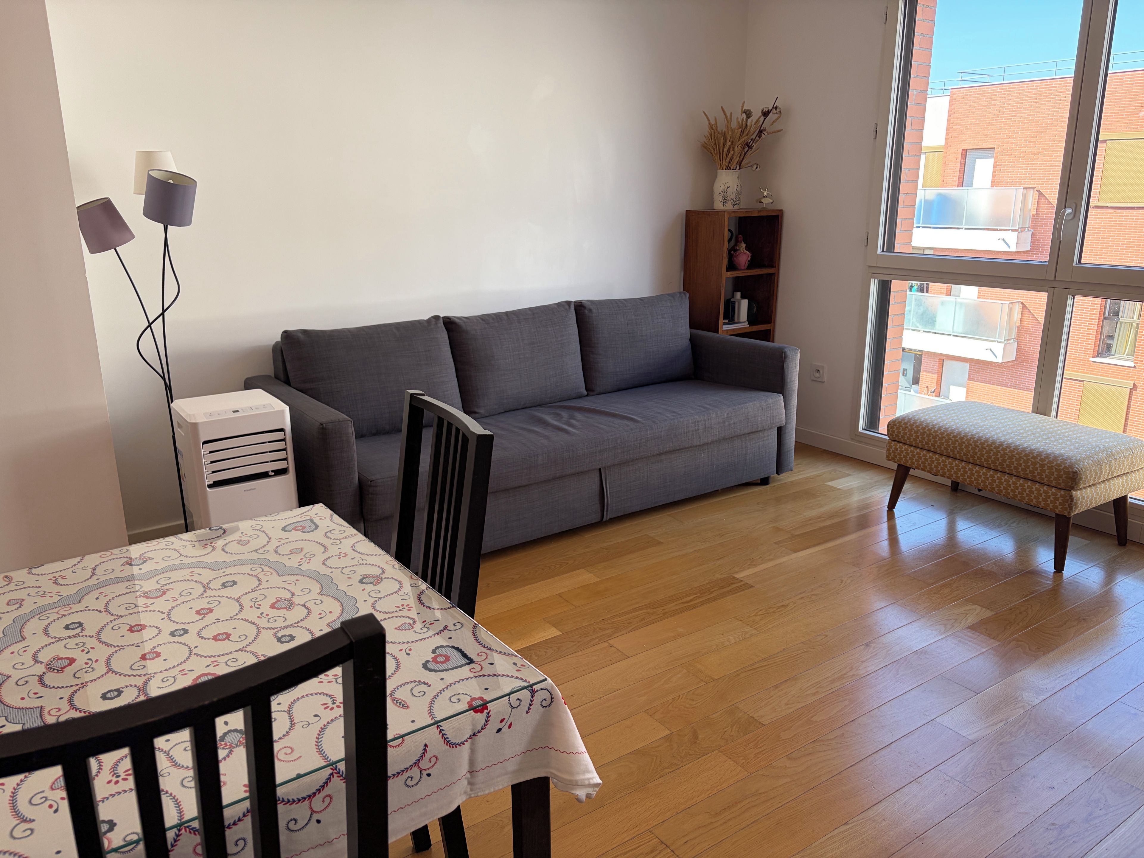 A living room with a grey sofa, a wooden table with a patterned tablecloth, a small bench, and a bookshelf.
