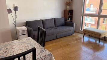 A living room with a grey sofa, a wooden table with a patterned tablecloth, a small bench, and a bookshelf.