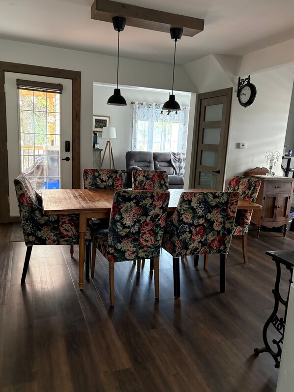A dining area with a wooden table and floral chairs, a black ceiling light, and a view into a living room.