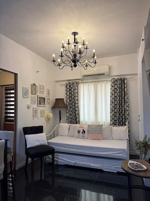 A bedroom with a bed, a chair, a table with a plate of food, a chandelier, and patterned curtains. -  1-bedroom condo with pool walking distance to NAIA Terminal 3 Pasay City (Pasay City)