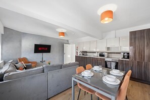 A modern living space with a grey sofa, a dining table set for four, and a kitchen area with wooden cabinets and a built-in oven. - Sleek Spacious 2BR Apartment in Liverpool (Liverpool)
