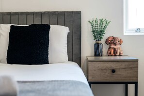 A bed with a black throw pillow and a white pillow, a wooden bedside table with a drawer, a small potted plant, and a wooden dog figurine. - Stylish 2BR Liverpool City Centre Apartment (Liverpool)