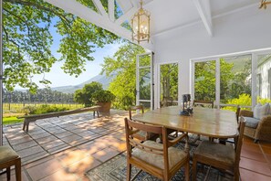 An outdoor patio with a wooden table and chairs, surrounded by large windows offering a view of a vineyard and hills. - Raithbury Hill House (Stellenbosch)