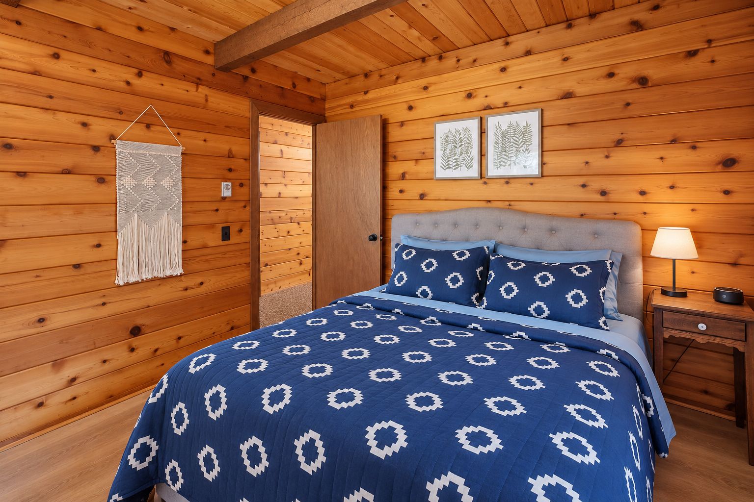 Een slaapkamer in een houten chalet met een bed, een nachtkastje, wanddecoratie van macramé en twee ingelijste foto's aan de muur.