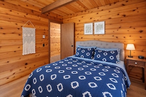 Een slaapkamer in een houten chalet met een bed, een nachtkastje, wanddecoratie van macramé en twee ingelijste foto's aan de muur.