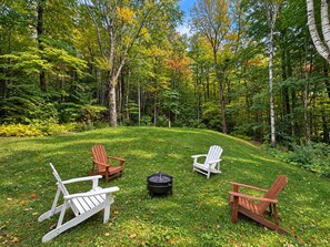 A grassy outdoor area with three Adirondack chairs and a fire pit.