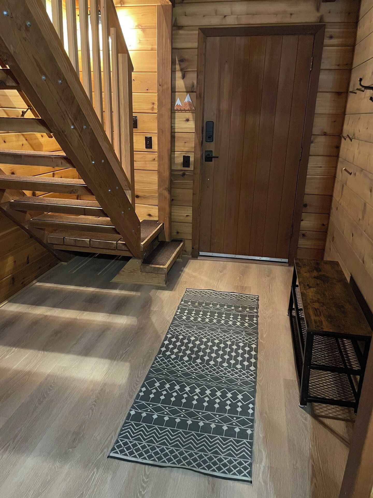 Eine hölzerne Treppe führt zu einer Tür mit einem Metallgriff, auf dem Boden liegt ein gemustertes Teppich und zur Rechten befindet sich eine Metallbank.